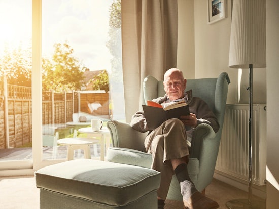 <p>Older man reading a book in his armchair at home [Source: iStock]</p>
