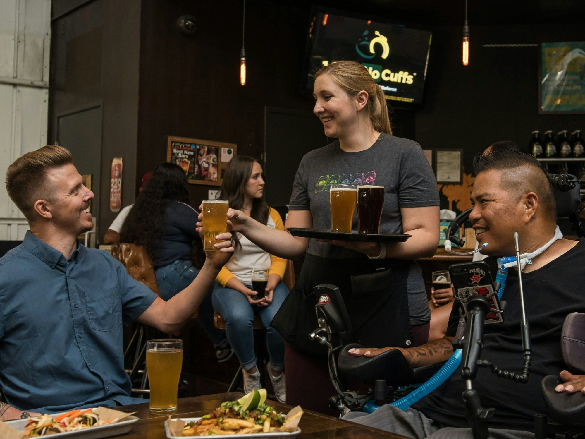 Two people being served beer at a pub table. One of them is using a wheelchair and other assistive technology support.