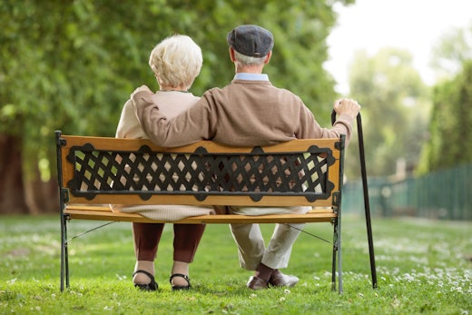 Two older people sit on a bench in a park