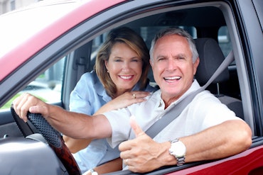 Two older people sit in a red car and look happy