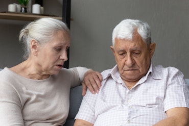 An older woman comforts her husband as he looks sad.