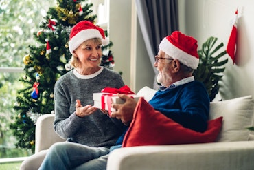 An older man gives a older woman a gift in front of a Christmas tree