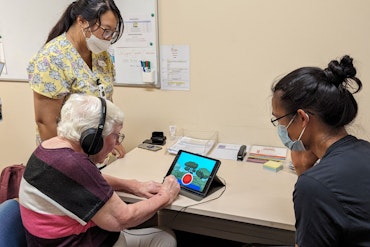 An older woman uses a touch-screen device while two women watch