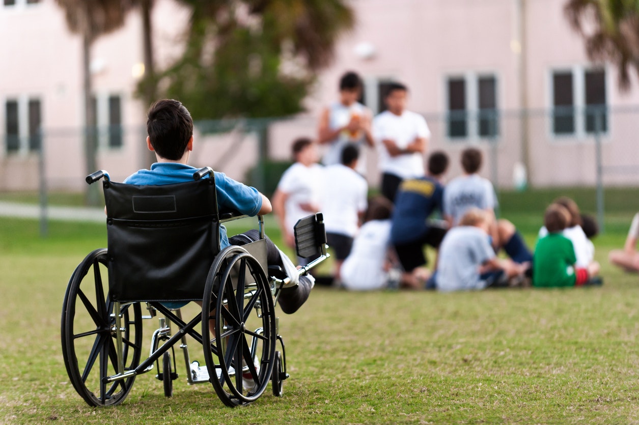 <p>twelve years old child sitting in a wheel chair watching other kids getting together in the park while he is left behind [Source Juanmonino, iStock]</p>
