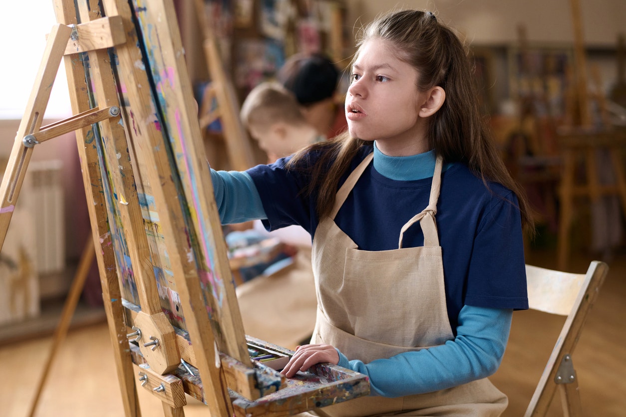 <p>Side view portrait of young girl with disability enjoying art class in studio and painting on canvas. [Source mediaphotos, istock]</p>
