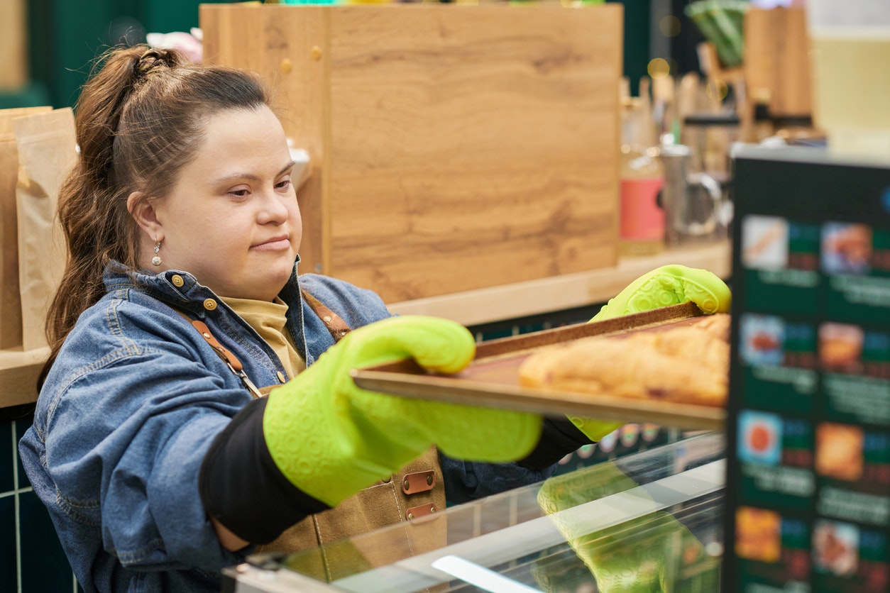 Portrait of young woman with Down syndrome working in bakery and pulling fresh croissants out of oven copy space