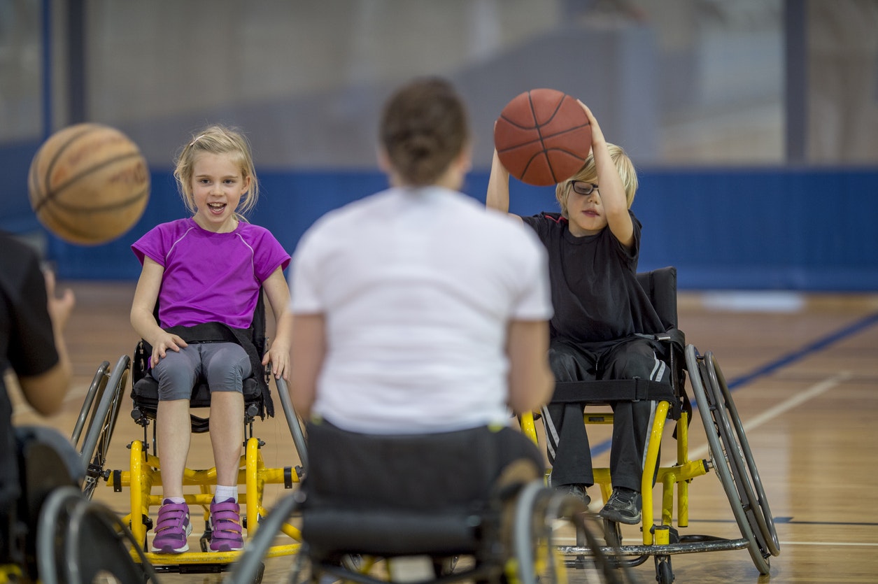 <p>Two children with disabilities are bouncing a basketball in the gym with their physical therapist. [Source FatCamera, iStock]</p>
