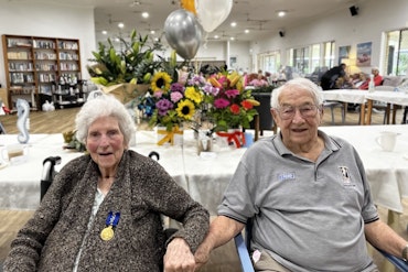 Two older people sit next to each other and hold hands