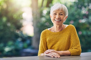 An older woman smiles at the camera, wearing a yellow jumper.