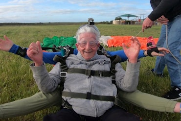 An older man wears skydiving gear on a field